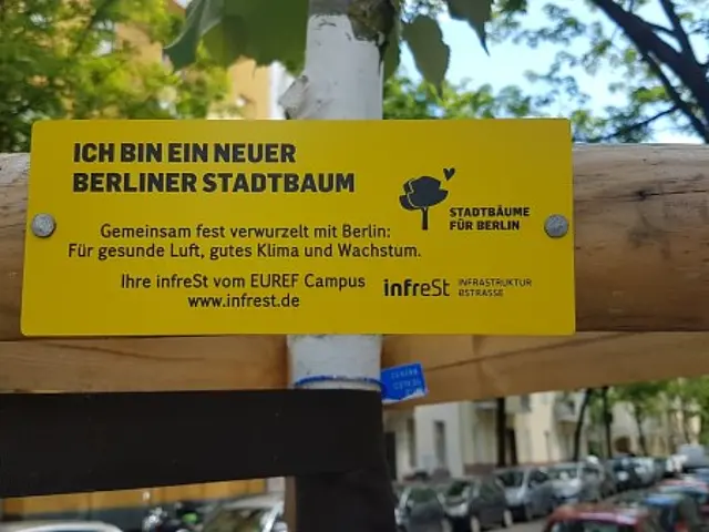 Gelbes Schild an einem jungen Stadtbaum mit Text zur Initiative Stadtbaeume fuer Berlin und Hinweis auf infrest vom EUREF Campus.