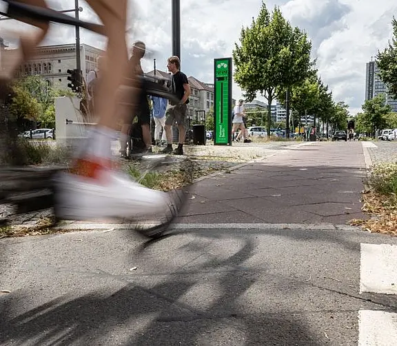 Schnell vorbeifahrendes Fahrrad im Vordergrund eines städtischen Radwegs; im Hintergrund stehen Personen, Bäume, Verkehr und moderne Gebäude.