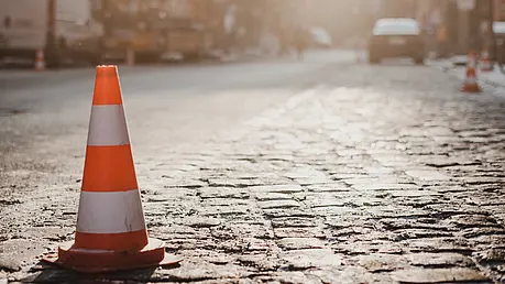 Ein orange‑weißer Verkehrskegel steht auf einer gepflasterten Straße im warmen Sonnenlicht, im Hintergrund unscharfe Baustellenszene.