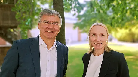 Andrea Pieper und Jürgen Besler in formeller Kleidung stehen nebeneinander im Freien; hinter ihnen Bäume, Wiese und ein helles Gebäude im Hintergrund.
