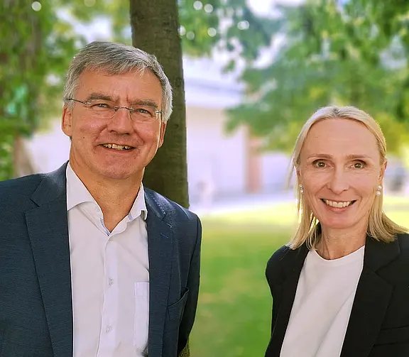 Andrea Pieper und Jürgen Besler in formeller Kleidung stehen nebeneinander im Freien; hinter ihnen Bäume, Wiese und ein helles Gebäude im Hintergrund.
