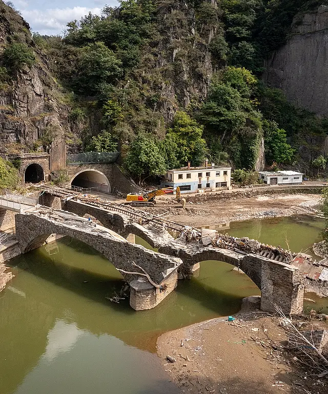 Zerstörte Steinbrücke in einer felsigen Flusslandschaft mit Tunneln und Gebäuden. Symbol für Infrastruktur‑Schäden und Wiederaufbau.